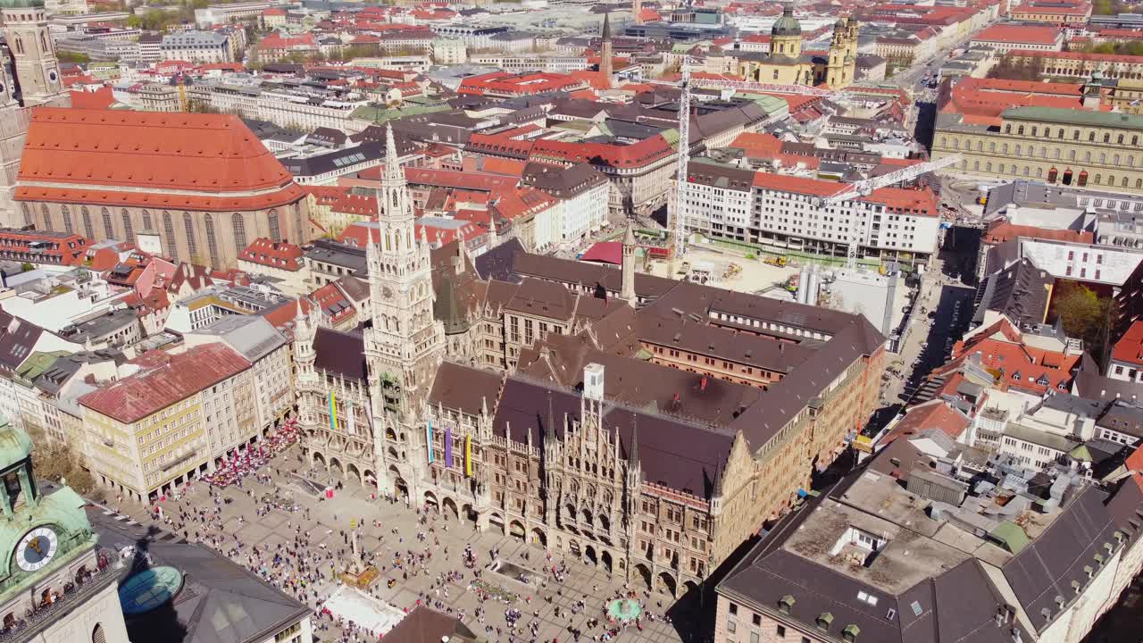 A drone orbits Marienplatz revealing Neues Rathaus, Frauenkirche, Peterskirche and Theatinerkirche among orange rooftops and busy square life under clear skies, drone orbiting shot