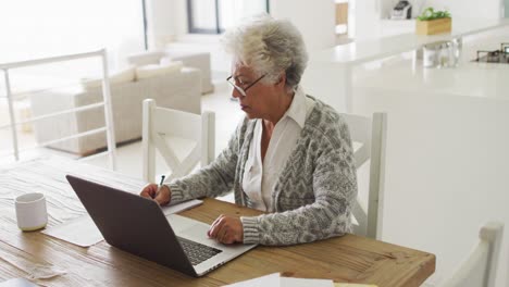 Thoughtful-african-american-senior-woman-taking-notes-and-using-laptop-at-home