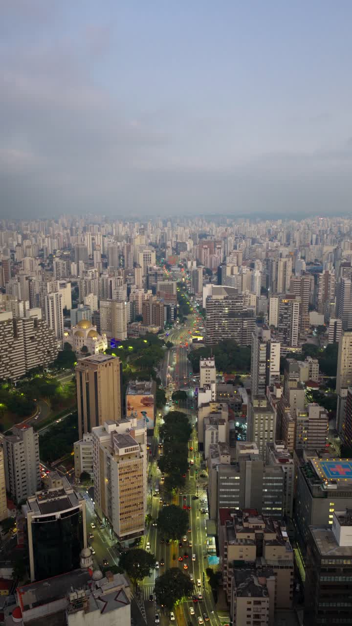 Aerial vertical zoom out of Av. Paulista showing the avenue stretching through São Paulo’s skyline at dusk with city lights and tall buildings.