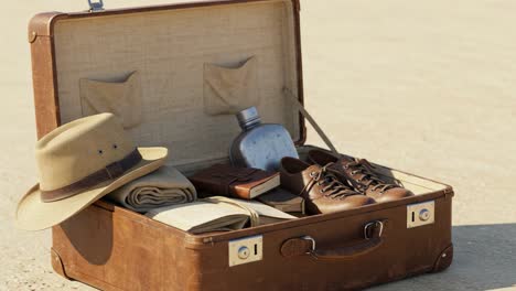 exploring the contents of an open suitcase featuring travel essentials like a hat, shoes, water flask, clothes, and a journal on a sandy terrain