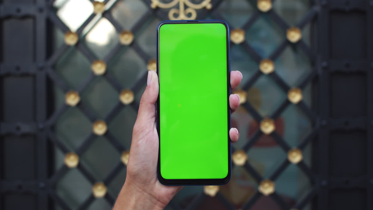 Hand holding a smartphone with a green screen in front of a decorative gate