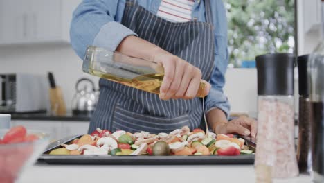 Sección-Media-De-Una-Mujer-Birracial-Vertiendo-Aceite-Sobre-Verduras-En-Una-Bandeja-Para-Hornear-En-La-Cocina,-Cámara-Lenta