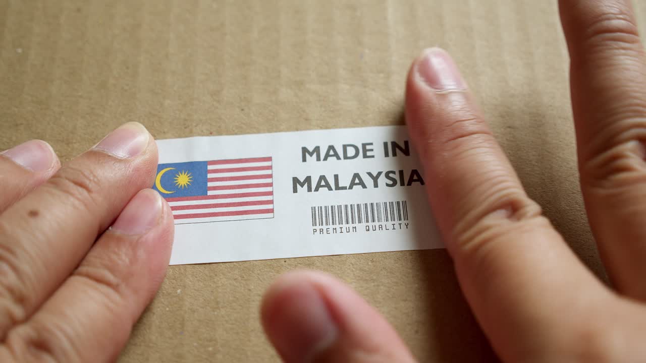 Hands applying MADE IN MALAYSIA flag label on a shipping box with product premium quality barcode