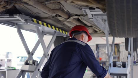 A-car-mechanic-in-helmet-standing-under-lifted-car,-making-notes-on-a-tablet