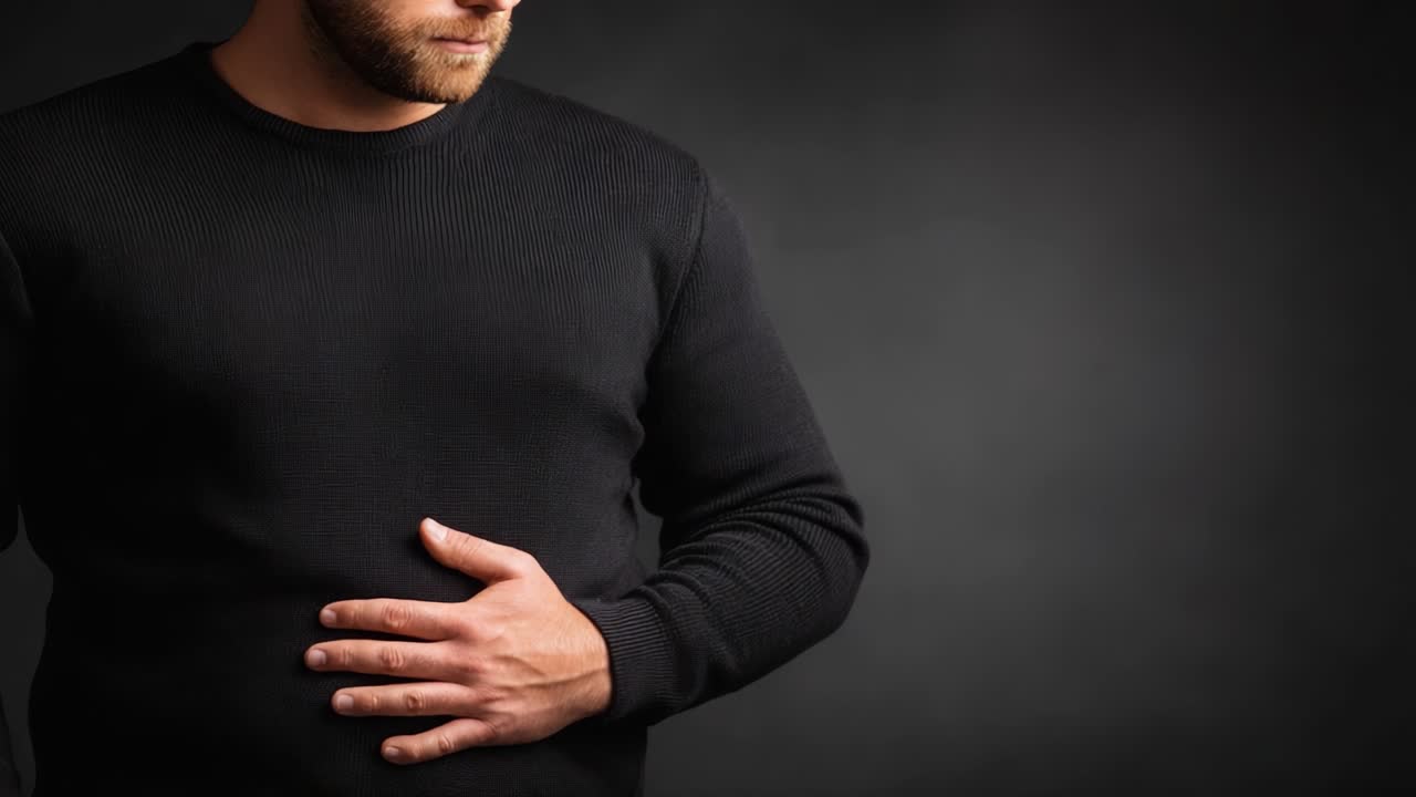 Ein Mann mit schmerzverzerrtem Gesichtsausdruck hält sich den Bauch, vermittelt Unbehagen und mögliche Schmerzen, während er vor einem dunklen, neutralen Hintergrund steht, der sein Leid hervorhebt.