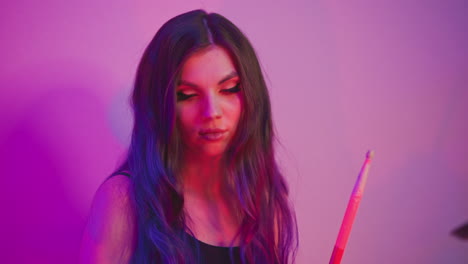 woman with long cascading hair bathed in neon purple and pink light holds drumstick calmly in hand while facing downward, creating moody studio ambiance with soft glow and artistic composition