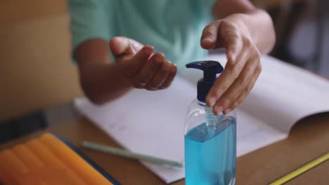 sección media de un niño desinfectando sus manos mientras está sentado en su escritorio en la escuela