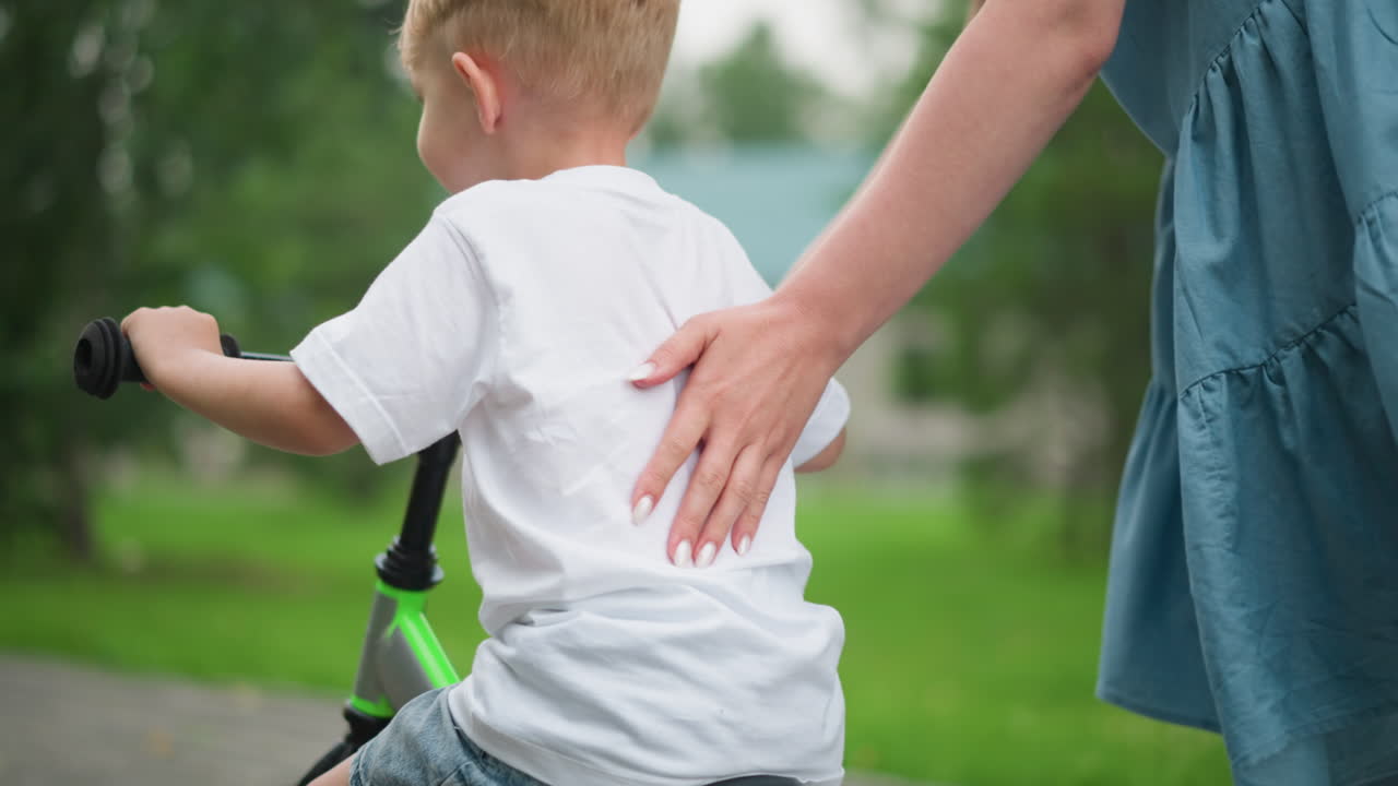 A woman with painted nails supports her young son who is learning how to ride a bicycle, the boy uses his leg for balance as she lets him move forward independently