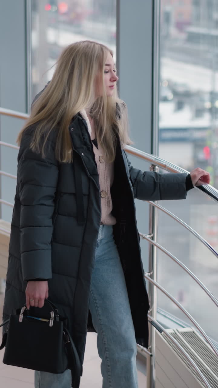 흑색 드백을 들고 철의 레일링 근처에 서 있는 여인, 뒷면에 도시 풍경과 사무실 건물이 있는 유리를 통해 바라보고, 바쁜 도시 환경에서 움직이는 사람들이 보인다