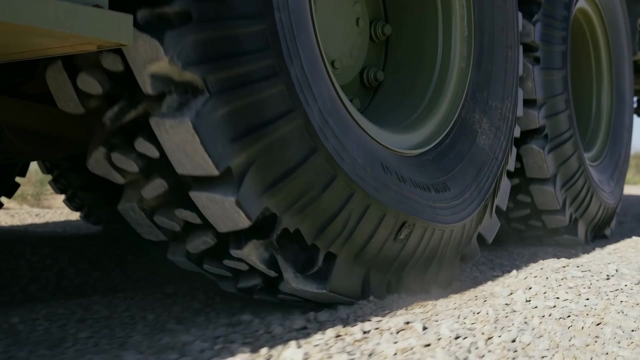 Low angle view of large off road vehicle wheels moving across a rough gravel surface, with sunlit open landscape in the background and dust and motion