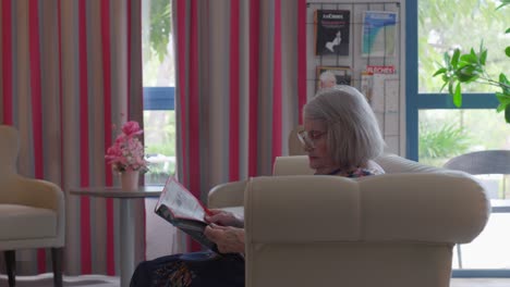 Elderly-woman-comfortably-seated-on-a-plush-sofa,-engrossed-in-reading-magazine