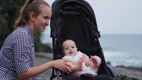 a young mother in this sweet family shares heartfelt moments with her baby in the stroller against the ocean backdrop, showcasing her nurturing and loving qualities