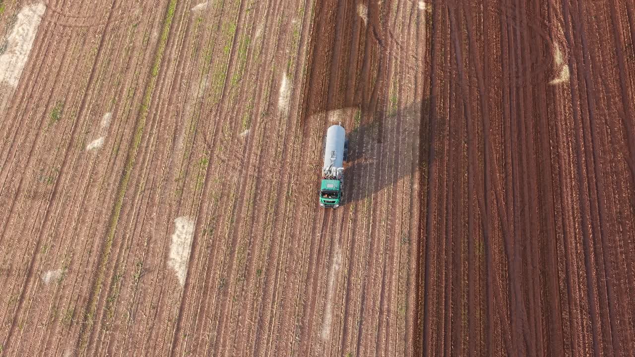 Aerial orbit of tractor spraying manure across farmland leaving darker soil trail behind