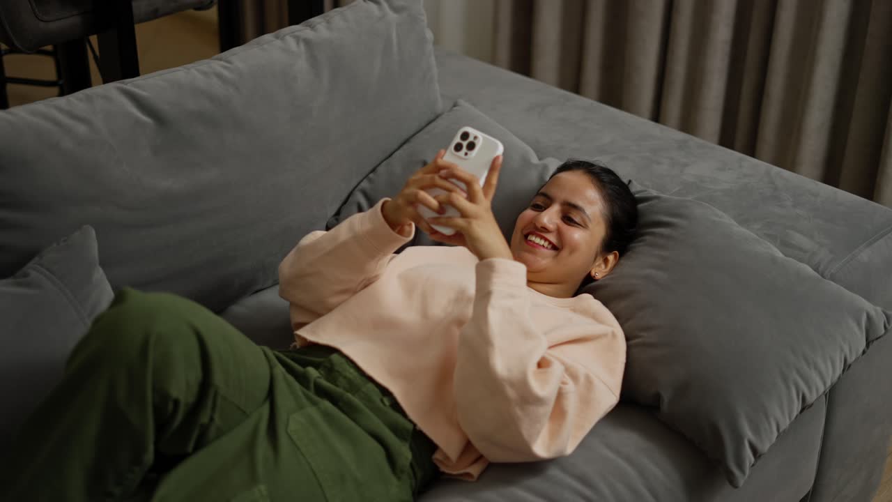 desde arriba, una feliz chica morena con un suéter beige y pantalones verdes se acuesta en una almohada en un sofá gris y se desplaza activamente por las redes sociales en su teléfono inteligente blanco durante el día