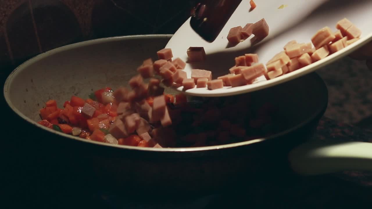 Aggiunta di prosciutto tagliato a cubetti in una padella con verdure saltate per preparare riso fritto. Scena di cucina casalinga spontanea