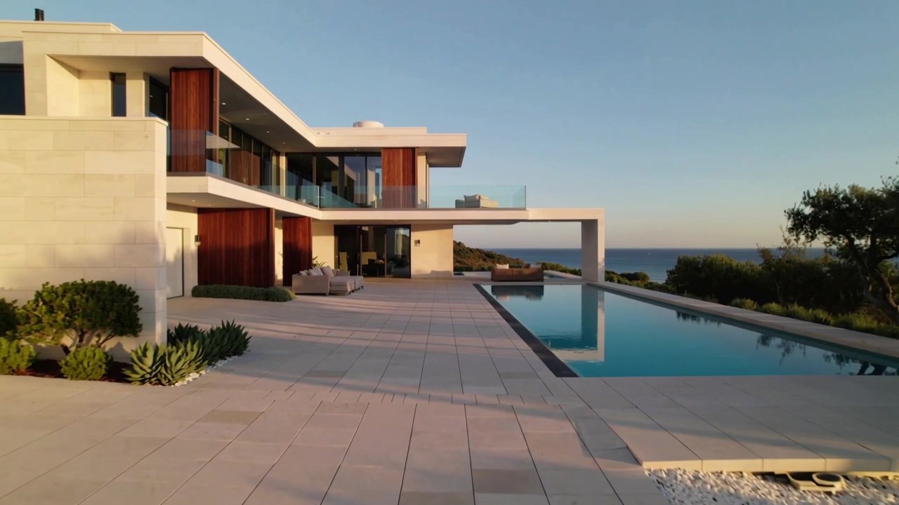 La telecamera panoramica a destra, rivelando una casa costiera con una lunga piscina e terrazza, che evidenzia la vista sul mare.