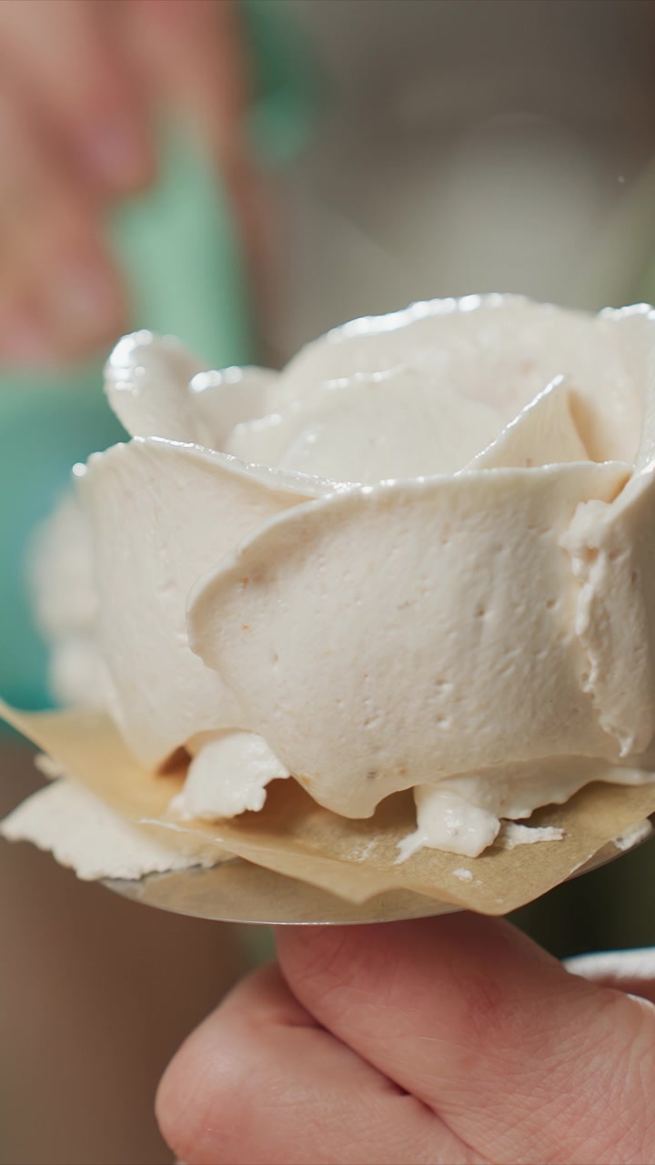 Close-up de mão segurando uma sobremesa de merengue em formato de rosa branca. A textura cremosa e os detalhes das pétalas são enfatizados. Uma pessoa vestindo verde é levemente visível em alguns quadros.