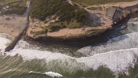 Toma-Aérea-Ascendente-De-Arriba-Hacia-Abajo-De-Gigantescos-Acantilados-Costeros-Con-Olas-Que-Llegan-Desde-El-Océano-Durante-La-Puesta-De-Sol---Mar-Del-Plata,-Argentina