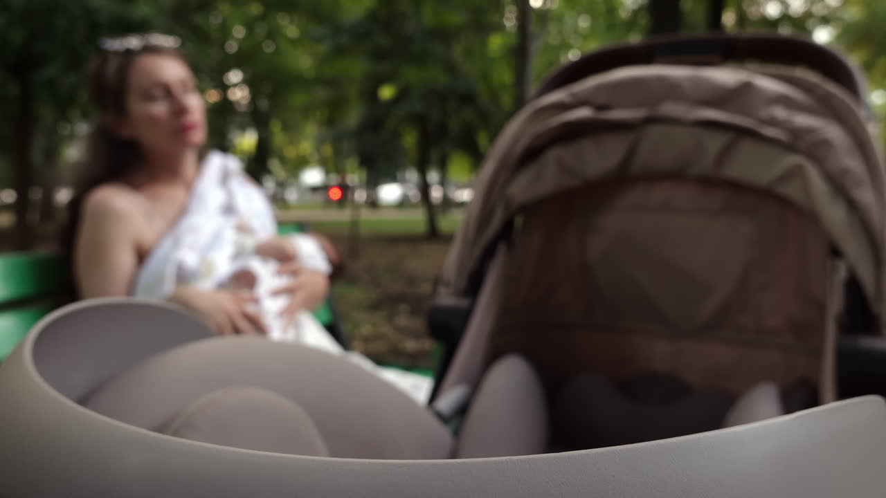 Close up of a stroller with a mother breastfeeding her baby while sitting on a park bench in the background