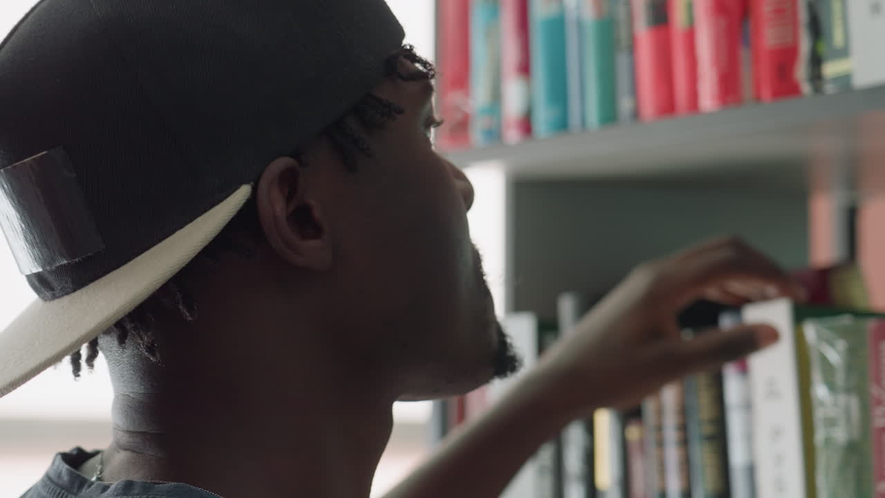 Side view of young man in cap moving hand across books on library shelf with focused expression, scanning titles attentively, showing curiosity, engagement, and determination in quiet indoor