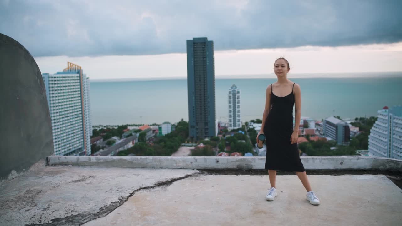 Shot of young woman in black dress standing on roof with sea view and looking front