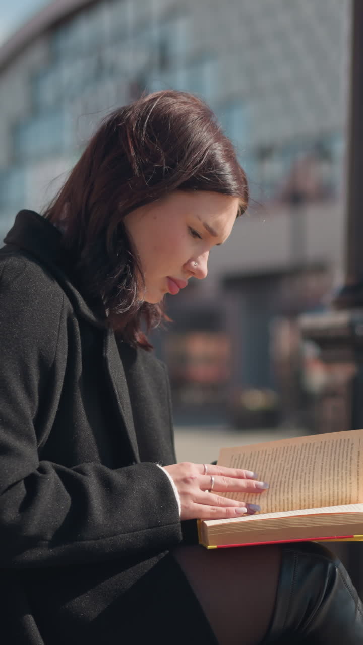 Young adult in black coat and leather boots sitting outdoors reading a book and flipping pages, with blurred street view and residential buildings in the background during bright sunny day