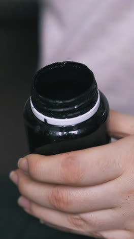 cu: experienced tailor opens small bottle with special leather paint holding over black cloth extreme close view