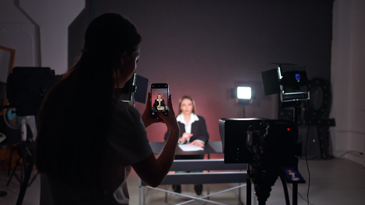 Rear view of a long-haired brunette taking photos on her phone. Girl stands among the video equipment in studio backstage.