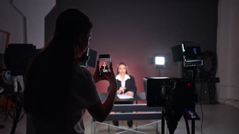 rear view of a long-haired brunette taking photos on her phone. girl stands among the video equipment in studio backstage.
