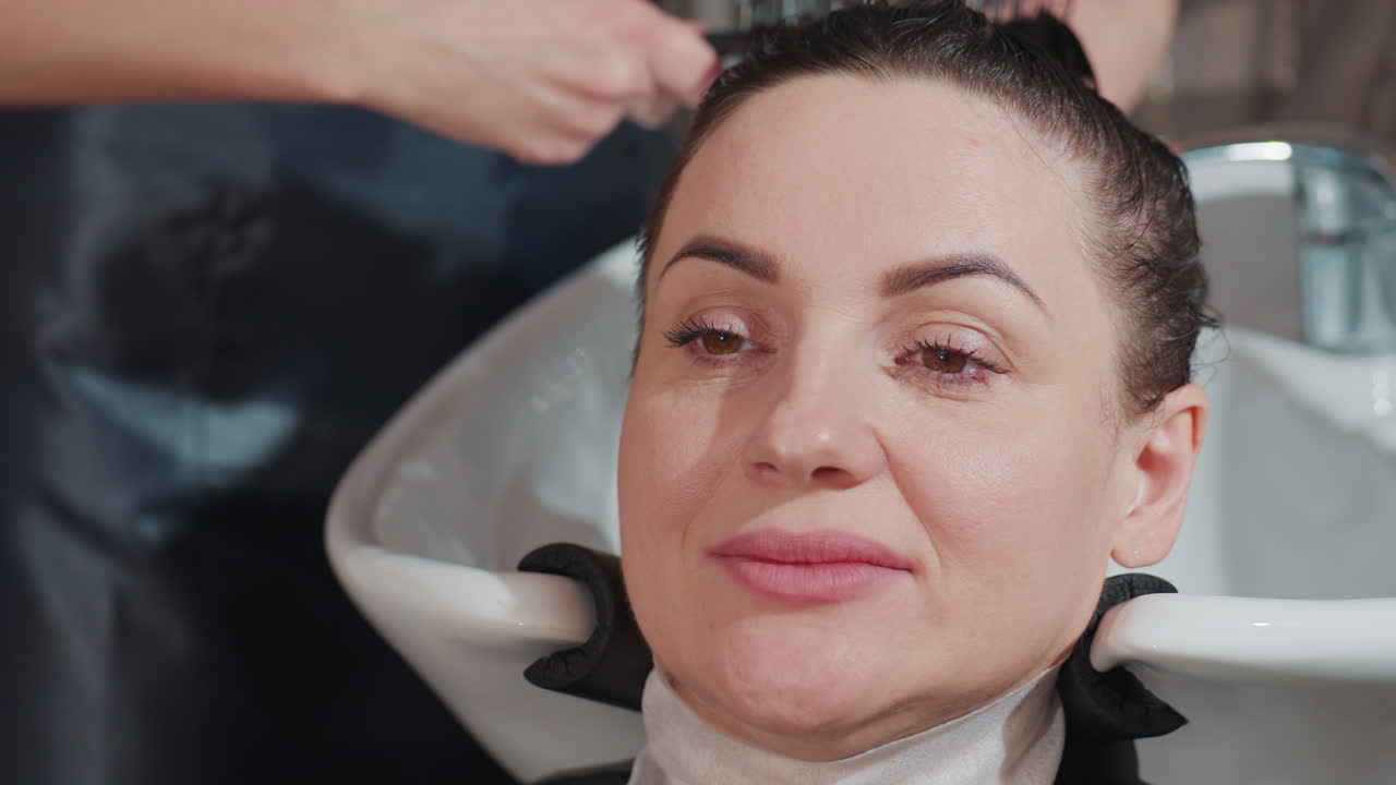 Eine kaukasische KundIn genießt eine beruhigende Wäsche, eine entspannte KundIn genießt ein sanftes Shampoo am Waschbecken des Salons, eine ruhige Salonumgebung, in der eine KundIn eine angenehme Haarwäsche mit warmem Wasser erhält