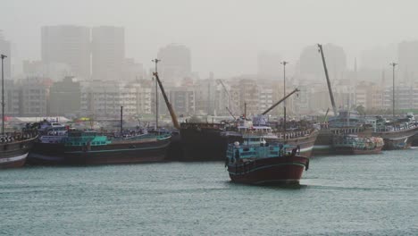 Dhow-Cargo-Boat-Sailing-At-Dubai-Creek-With-City-Covered-In-Fog-In-The-Morning-In-Dubai,-UAE