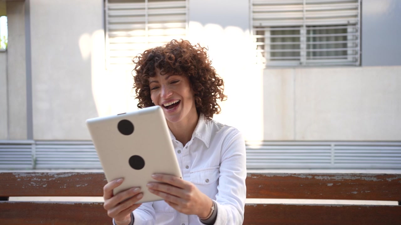 A young woman is sitting on a bench in an outdoor park using her tablet while enjoying the sun