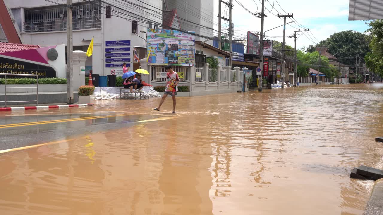 A Thai man approaches a dry area after traversing flooded streets near three girls of Chiang Mai Christian School seated on stairs with umbrellas Sandbags are visible blocking the water September 2024