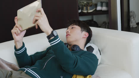 asian boy playing games on digital tablet lying on the couch at home