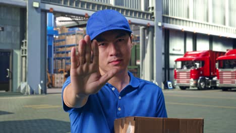 close up of asian male courier in blue uniform making stop gesture and shaking his head while delivering a carton, outside of logistics distributions warehouse
