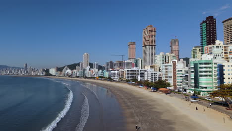 drone traces waves along the shore that fade into backdrop of city buildings and hills. ideal for coastal tourism or environmental storytelling.itapema - meia praia, brazil
