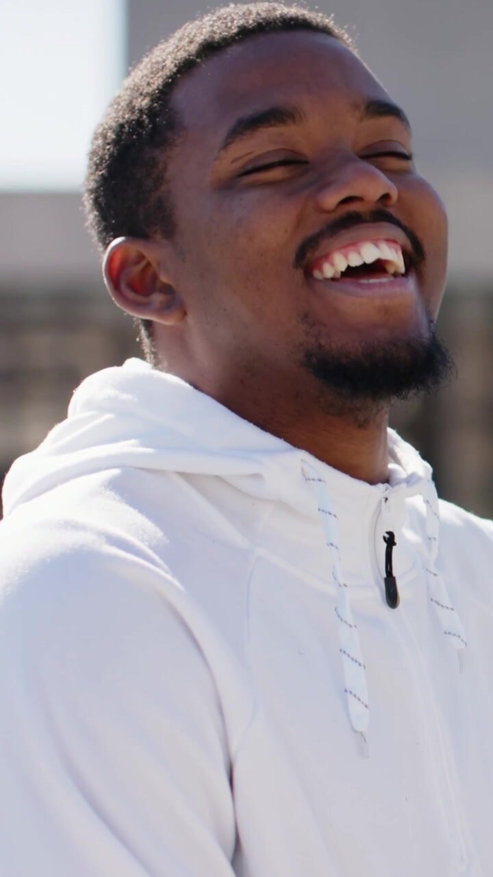 Vertical video: Smiling man shifting into laughter on rooftop, with white hoodie and blurred facade