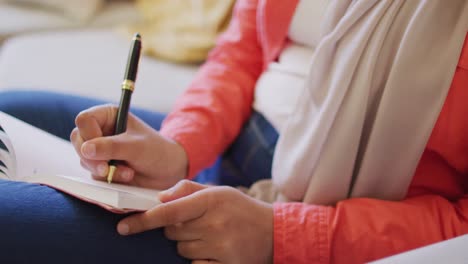 Video-of-midsection-of-biracial-woman-writing-with-pen-in-notebook-sitting-in-living-room-at-home