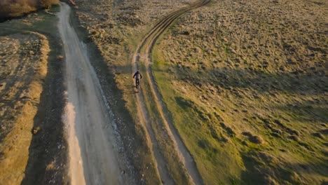 cyclist rides down a dirt path through grassy hills in warm sunset light, aerial view