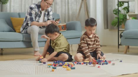 full body of asian kids assemble the construction set colorful plastic toy brick, the father sitting on a sofa using smartphone