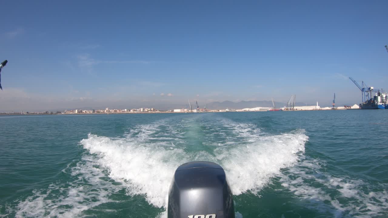 항구에서 멀어져가는 배의 후미, 크레인과 건물은 수평선에서 멀리 떨어져 있고 엔진은 해수에서 웨이크를 그립니다.