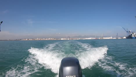 detrás de un barco que se aleja del puerto, las grúas y los edificios están lejos en el horizonte mientras el motor dibuja una estela en el agua del mar