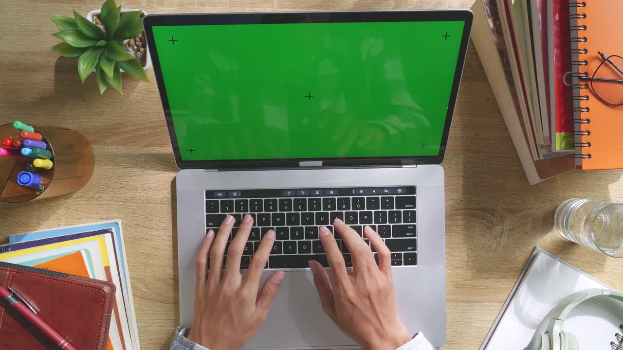 Top down view of a laptop computer with mock up green screen chromakey display on a wooden office desk next to notebook with pens, glasses, and a glass of water. Slow zoom out, close up