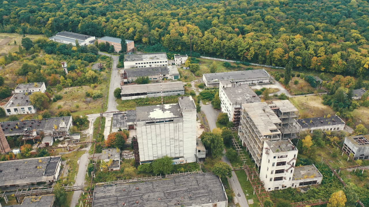 Empty factory constructions. Flight over abandoned buildings with old roofs. Ruined industrial territory near the forest.