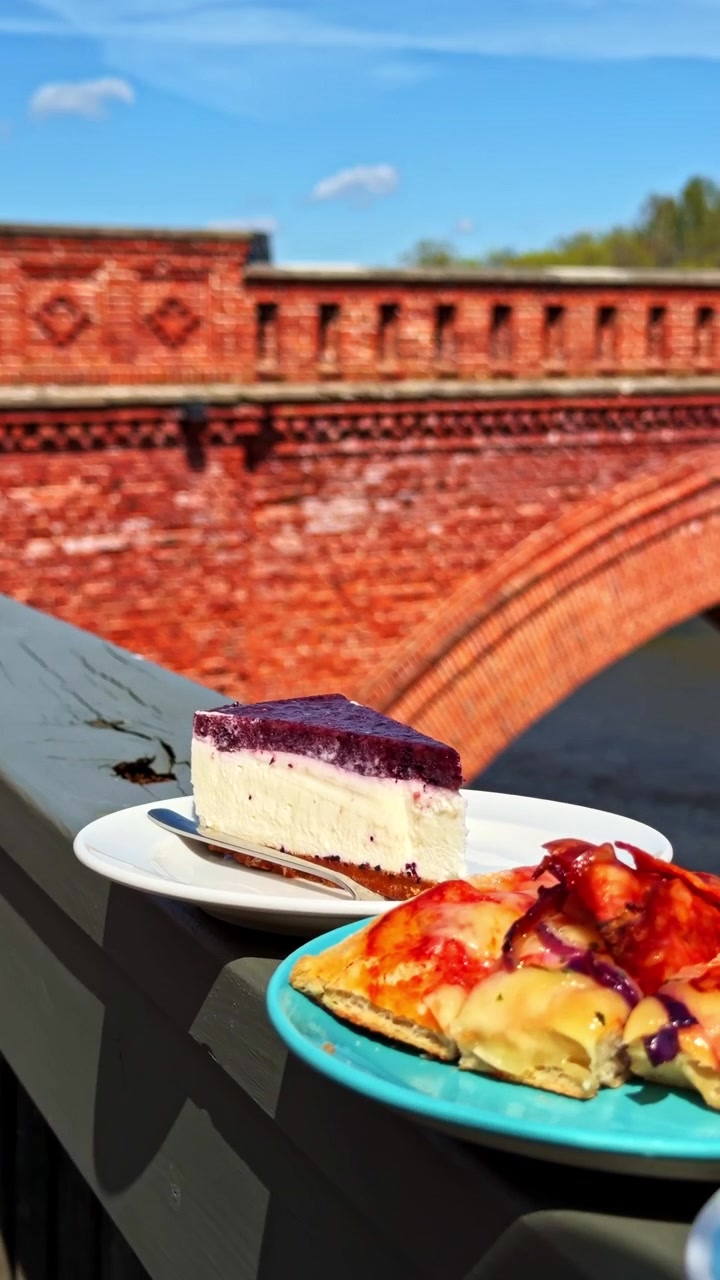 Fågel sitter på träräcke vid röd tegelbro med cheesecake och paj i närheten.