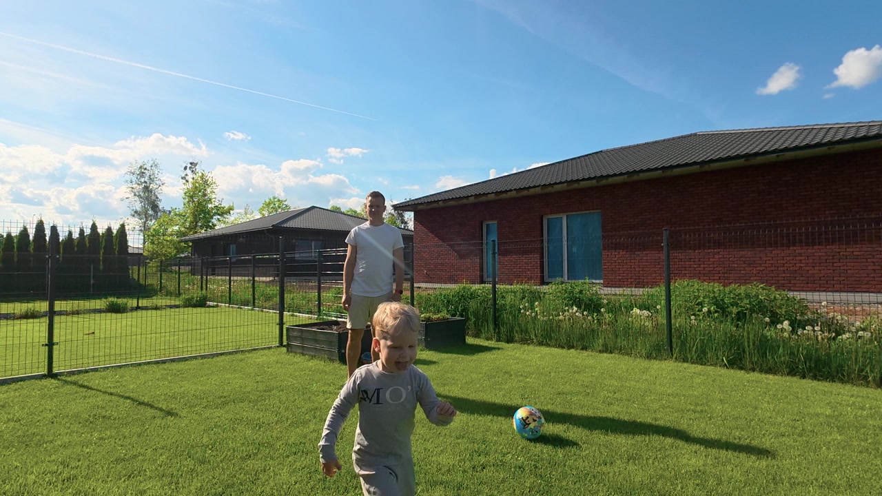 A dad and young boy play with a ball and frisbee in a fenced backyard on a sunny afternoon. Vibrant green grass, casual attire, clear blue sky, and soft natural lighting create a cheerful, family-frie