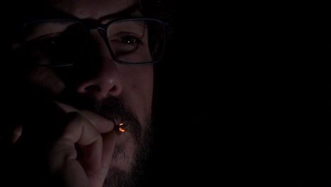 young adult male with glasses and beard smoking cigarette in dark room reflecting digital screen