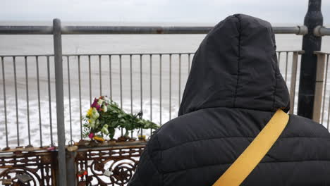 a pensive lonely sad mourning person in a hooded jacket stands by a seaside railing, observing a bouquet of flowers placed in remembrance of loss on bereavement