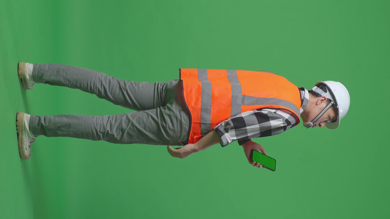 Full Body Back View Of Asian Male Engineer With Safety Helmet Working On A Green Screen Smartphone And Looking Around While Standing In The Green Screen Background Studio