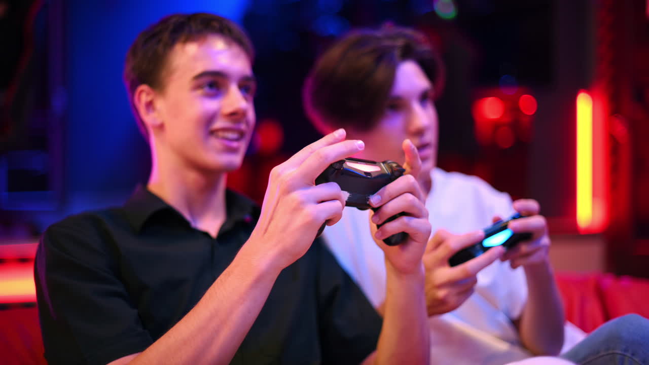 Dva dospívající kamarádi hrají na herní konzoli pomocí gamepadů, baví se a usmívají se. Modré a červené osvětlení